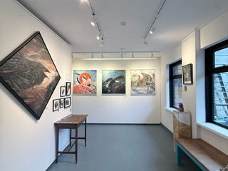 An art gallery interior with multiple framed paintings displayed on white walls, including a large dark bird painting on the left and three smaller paintings in the center. A wooden table and bench are placed in the gallery space, with windows and track lighting visible.