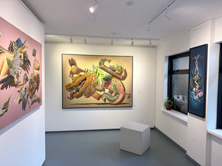 A large framed art print depicting a colorful, stylized crocodile in green, gold, and pink tones, displayed on a white gallery wall in a modern art studio with other paintings visible.